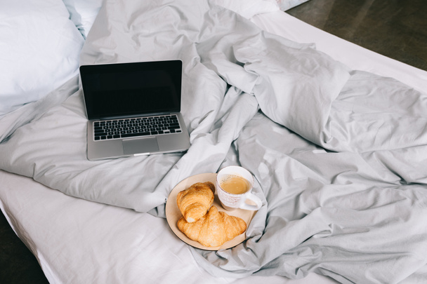 taza de café y croissants en el plato y portátil abierto en la cama
 - Foto, Imagen