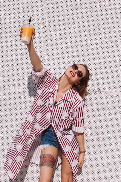 hermosa mujer joven sosteniendo cóctel en taza de plástico en frente de la pared blanca
 - Foto, Imagen