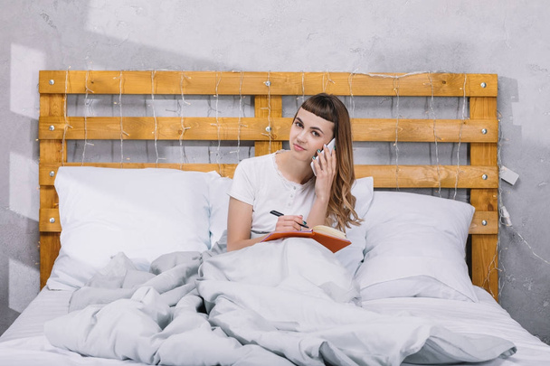 Uśmiechnięta dziewczyna coś pisze notebook i rozmawiać przez smartfon w łóżku - Zdjęcie, obraz