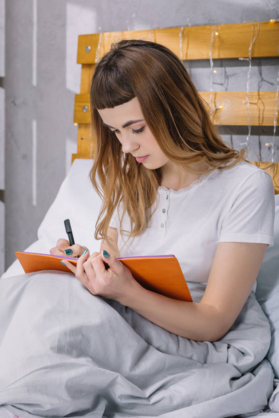 το κορίτσι γράφει κάτι στο σημειωματάριο στο κρεβάτι πρωί - Φωτογραφία, εικόνα