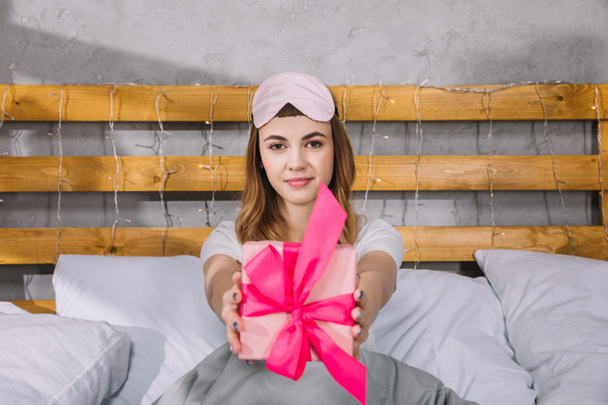 щаслива дівчина сидить у ліжку і показує подарункову коробку
 - Фото, зображення