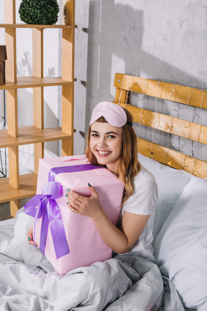 щаслива дівчина сидить у ліжку з подарунковою коробкою і дивиться на камеру
 - Фото, зображення