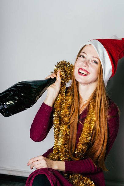 redhead happy girl in a red cap like Santa Claus celebrates the new year, holding a bottle of champagne - Foto, afbeelding