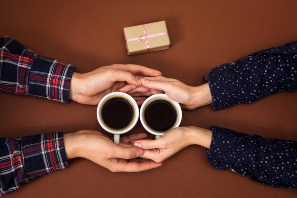 Чоловік і жінка тримають чашки з кавою в руках. Жіночі та чоловічі руки закриті на коричневому тлі. Хлопець дарує подарунок дівчині
. - Фото, зображення