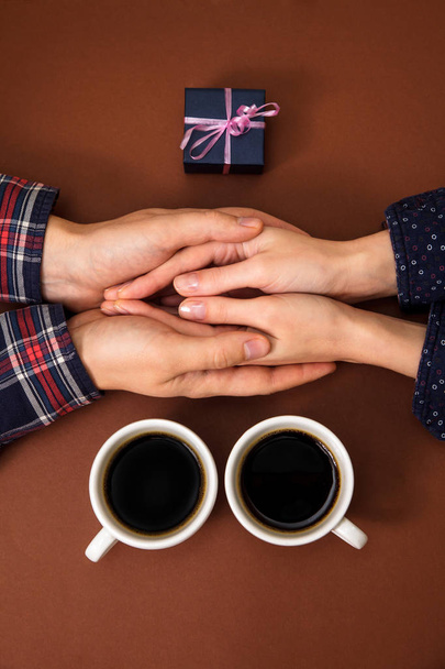 Мужчина и женщина держат чашки с кофе в руках. Женские и мужские руки закрыты на коричневом фоне. Парень делает подарок девушке.
. - Фото, изображение