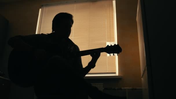Тёмный силуэт молодого привлекательного мужчины-музыканта сочиняет музыку на гитаре и играет, другой музыкальный инструмент на переднем плане, размытый
 - Кадры, видео