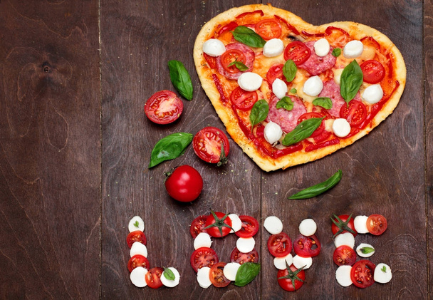 Pizza in shape heart with salami, tomatoes, basil and mozzarella with knife for pizza.Against a dark wooden brown background. Inscription love of mozzarella tomatoes and basil .Top View. Creative vale - Photo, Image