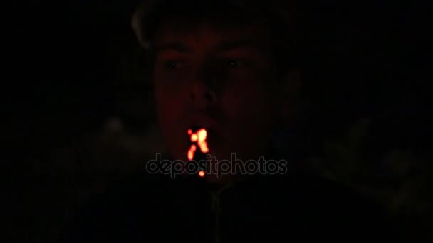 Face of boy blowing at smoldering branch at dark night - Filmati, video
