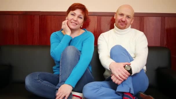 Middle-aged couple sit and smile on black sofa indoor - Imágenes, Vídeo