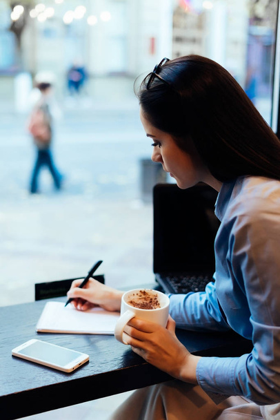 young girl sitting at a table drinking coffee and writing in a notebook - Foto, immagini