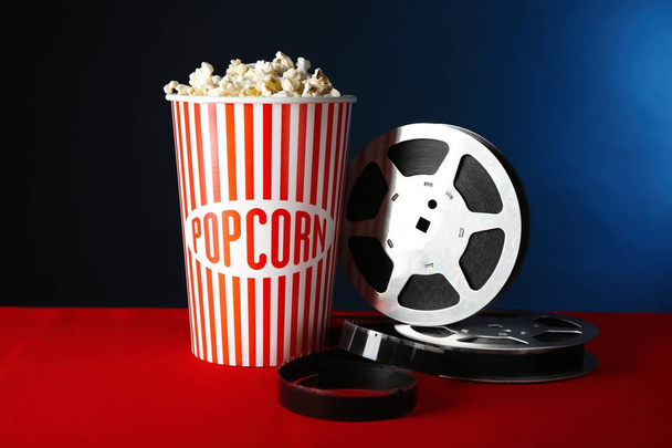Paper cup with tasty popcorn and movie reel on table against dark background - Foto, Imagen