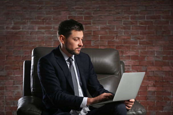 Handsome businessman with laptop sitting in comfortable armchair on brick wall background - Foto, afbeelding