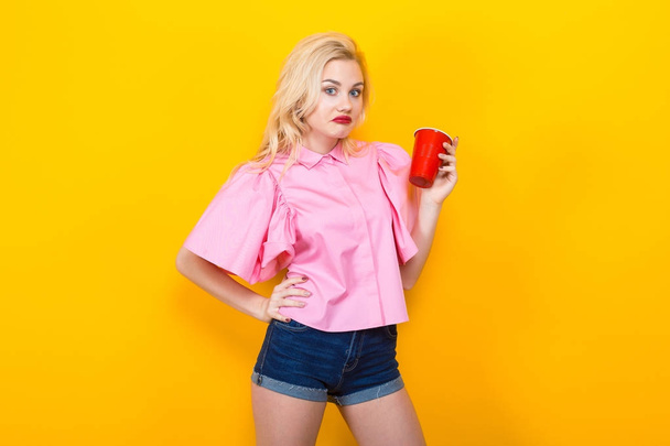stylish young woman in pink blouse posing with red plastic cup on yellow background - Fotoğraf, Görsel
