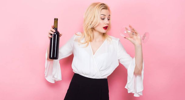 Όμορφη νεαρή γυναίκα με το άδειο ποτήρι και ένα μπουκάλι κόκκινο κρασί σε ροζ φόντο - Φωτογραφία, εικόνα