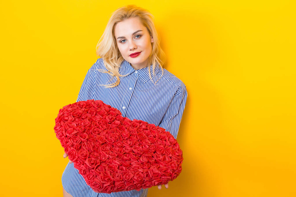 Mooie jonge vrouw in blauw shirt houden grote rode hart gemaakt van rozen op gele achtergrond - Foto, afbeelding