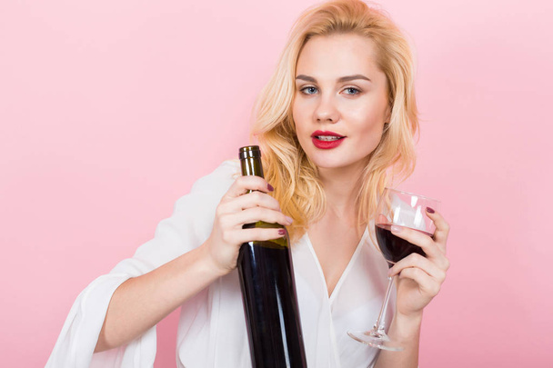 Όμορφη νεαρή γυναίκα με γυαλί και μπουκάλι κόκκινο κρασί σε ροζ φόντο - Φωτογραφία, εικόνα