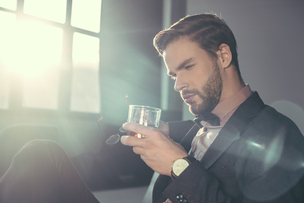 handsome bearded young man holding eyeglasses and glass of whisky - Fotó, kép