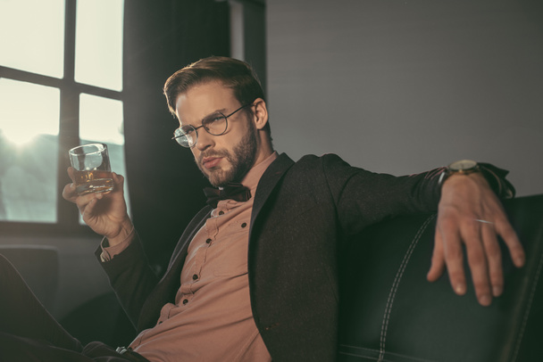 pohledný elegantní mladý muž v brýlích drží sklenici whisky a při pohledu na fotoaparát - Fotografie, Obrázek