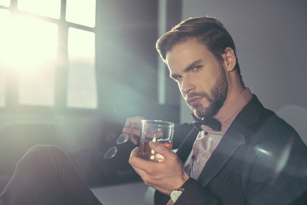 stylish young man holding eyeglasses and glass of whisky, looking at camera - Φωτογραφία, εικόνα