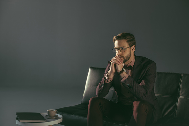 zamyšlený elegantní mladík v brýle sedí na gauči a koukal - Fotografie, Obrázek