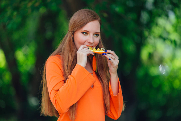 Portrait of pretty red hair woman with juicy delicious orange at summer green park. - Valokuva, kuva