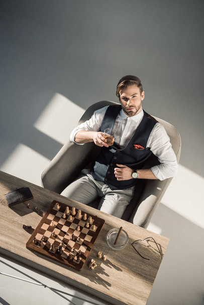 high angle view of young businessman looking at camera while drinking whisky and playing chess  - Photo, Image