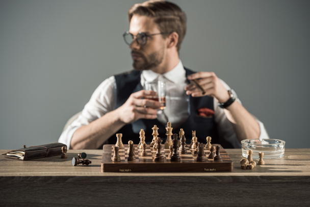 vista de cerca del tablero de ajedrez con figuras y el hombre bebiendo whisky y fumando cigarro detrás
 - Foto, Imagen