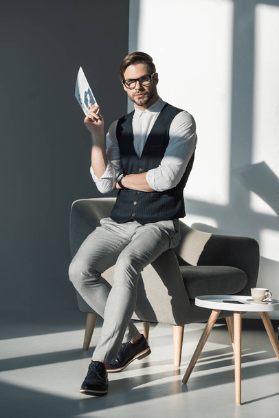 full length view of stylish young businessman holding newspaper and looking at camera - Valokuva, kuva