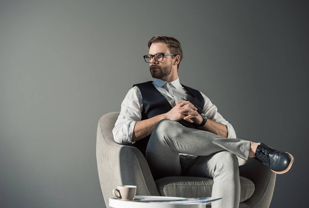 portrait of handsome young businessman in eyeglasses looking away while sitting in armchair isolated on grey - Foto, immagini