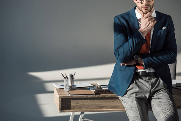обрезанный снимок стильного молодого бизнесмена с рукой в подбородке, сидящего на столе
 - Фото, изображение