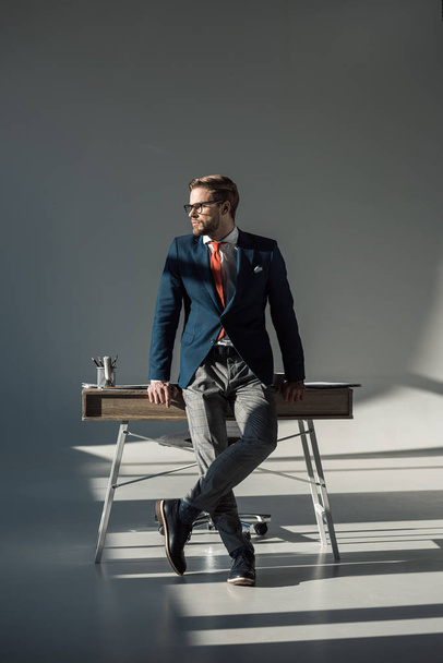 stylish young businessman in formal wear sitting on table and looking away - 写真・画像