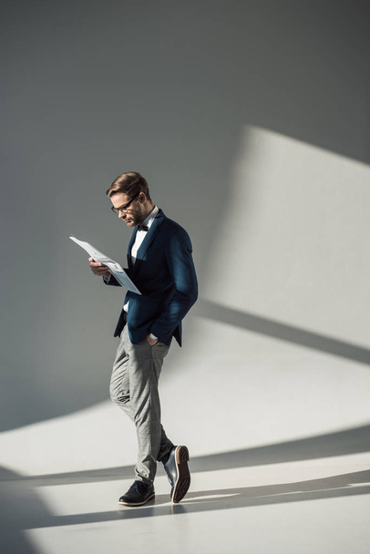 В полный рост красивый стильный бизнесмен в очках читает газету на сером
 - Фото, изображение