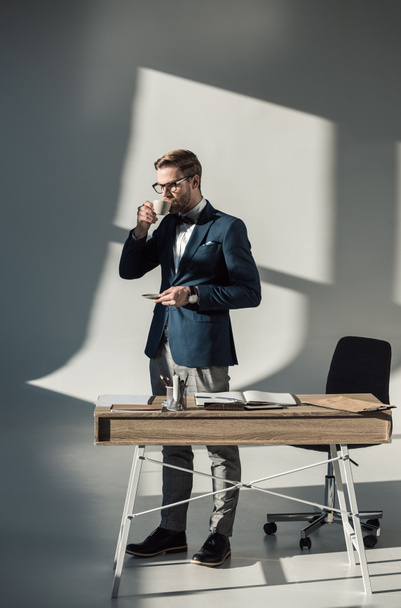 handsome young businessman in eyeglasses drinking coffee at workplace - Фото, изображение