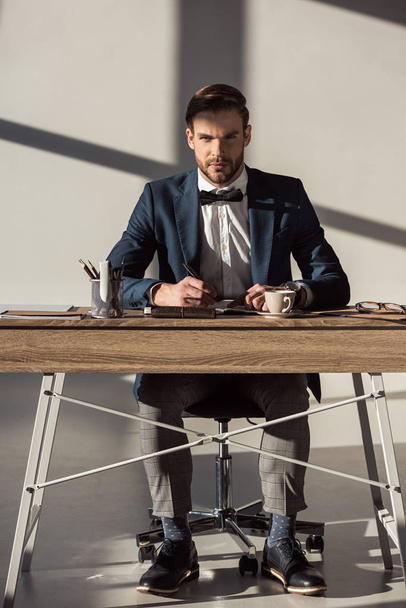 vista de longitud completa de hombre de negocios elegante guapo mirando a la cámara mientras escribe en el diario en el lugar de trabajo
 - Foto, Imagen