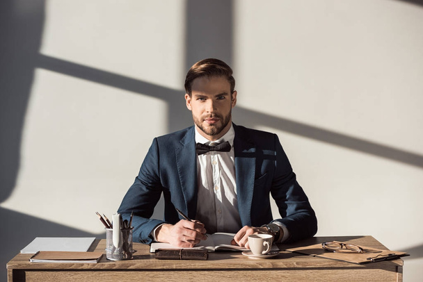 knappe stijlvolle zakenman camera kijken tijdens het schrijven in dagboek op werkplek - Foto, afbeelding