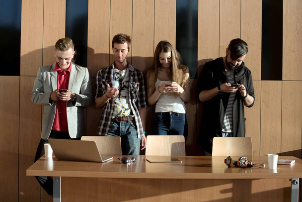 Four friends standing near the wooden wall with the phones in their hands. Casual lifestyle. - Foto, Imagen