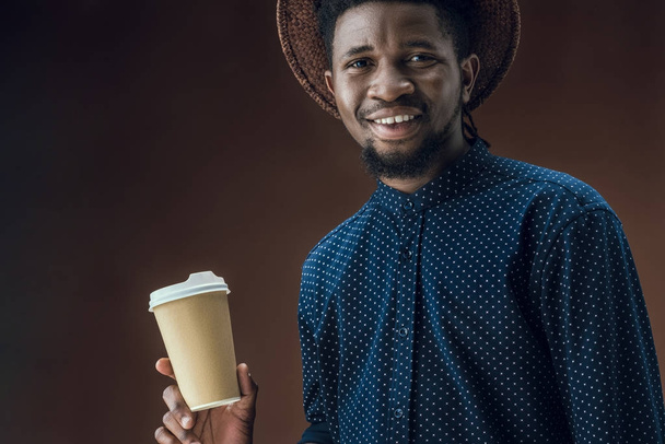 lachende Afro-Amerikaanse man met wegwerp koffiekopje geïsoleerd op brown - Foto, afbeelding