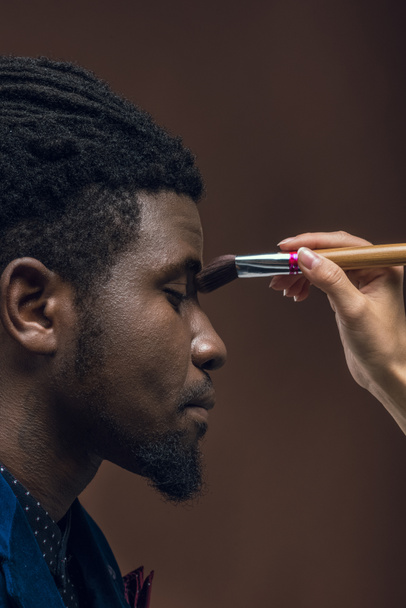 immagine ritagliata di donna che applica fondotinta in polvere con pennello per il trucco sul viso dell'uomo afroamericano isolato sul marrone
 - Foto, immagini