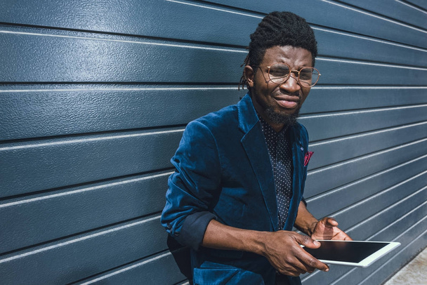 stílusos afrikai amerikai férfi, a gazdaság tabletta és látszó-on fényképezőgép kék kabát - Fotó, kép