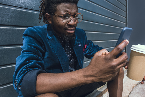 stylish african american man in blue jacket holding coffee to go and using smartphone - Photo, Image