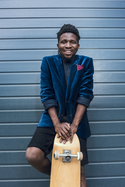 усміхнений стильний афроамериканський чоловік у блакитній куртці, що спирається на скейтборд
 - Фото, зображення