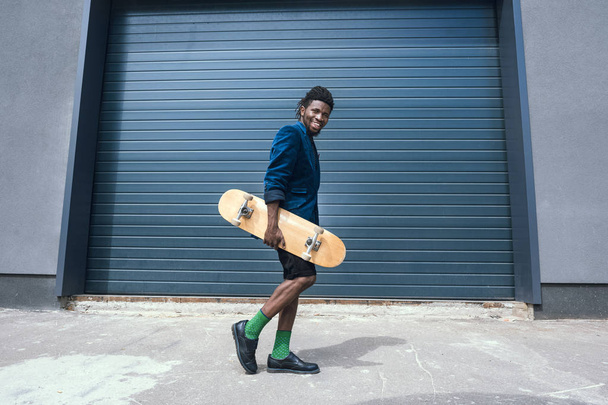 stijlvolle Afro-Amerikaanse man in blauwe jas met skateboard lopen op straat - Foto, afbeelding