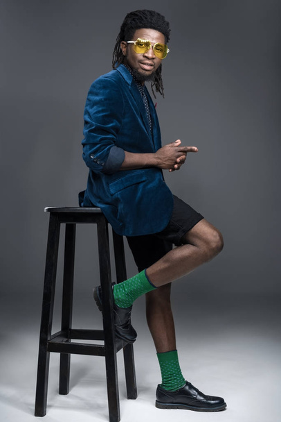 elegante uomo afroamericano in giacca blu appoggiato sulla sedia su grigio
  - Foto, immagini