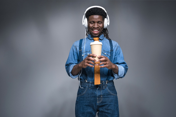 happy african american man listening music and holding coffee to go on gray - 写真・画像