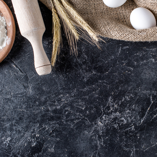 flat lay with wheat, raw eggs on sack cloth and wooden rolling pin on dark marble surface - Photo, Image