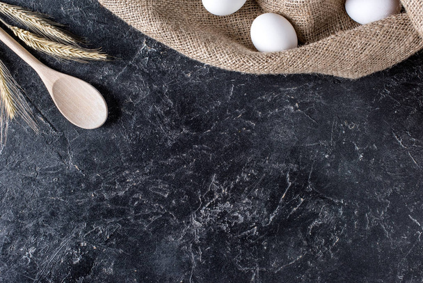 flache Lage mit Weizen, rohen Eiern auf Sacktuch und Holzlöffel auf dunkler Marmoroberfläche - Foto, Bild