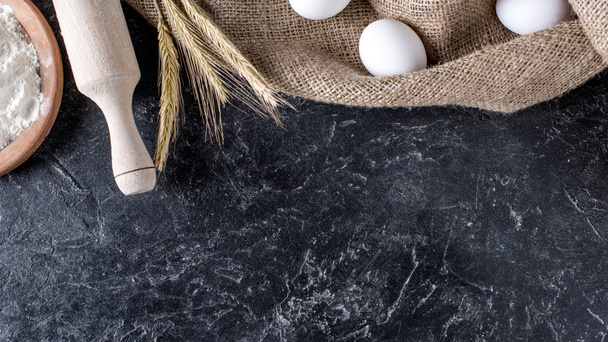 flat lay with wheat, raw eggs on sack cloth and wooden rolling pin on dark marble surface - Photo, Image