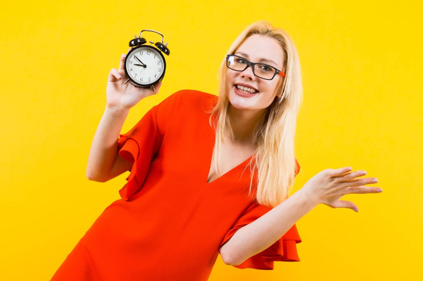 Mujer joven emocional en vestido rojo sosteniendo reloj despertador sobre fondo amarillo
 - Foto, Imagen