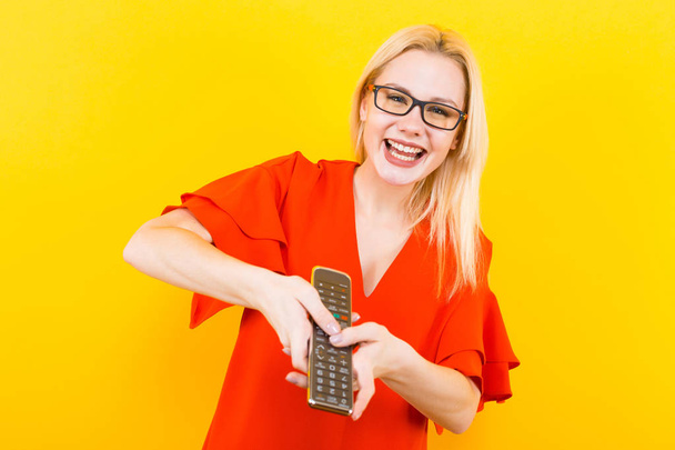 Beautiful young woman in red dress posing with remote control on yellow background - Fotografie, Obrázek