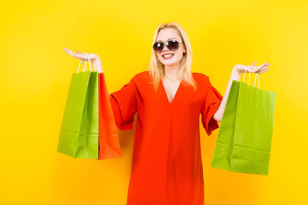Beautiful young woman in red dress posing with colorful shopping bags on yellow background - Фото, изображение